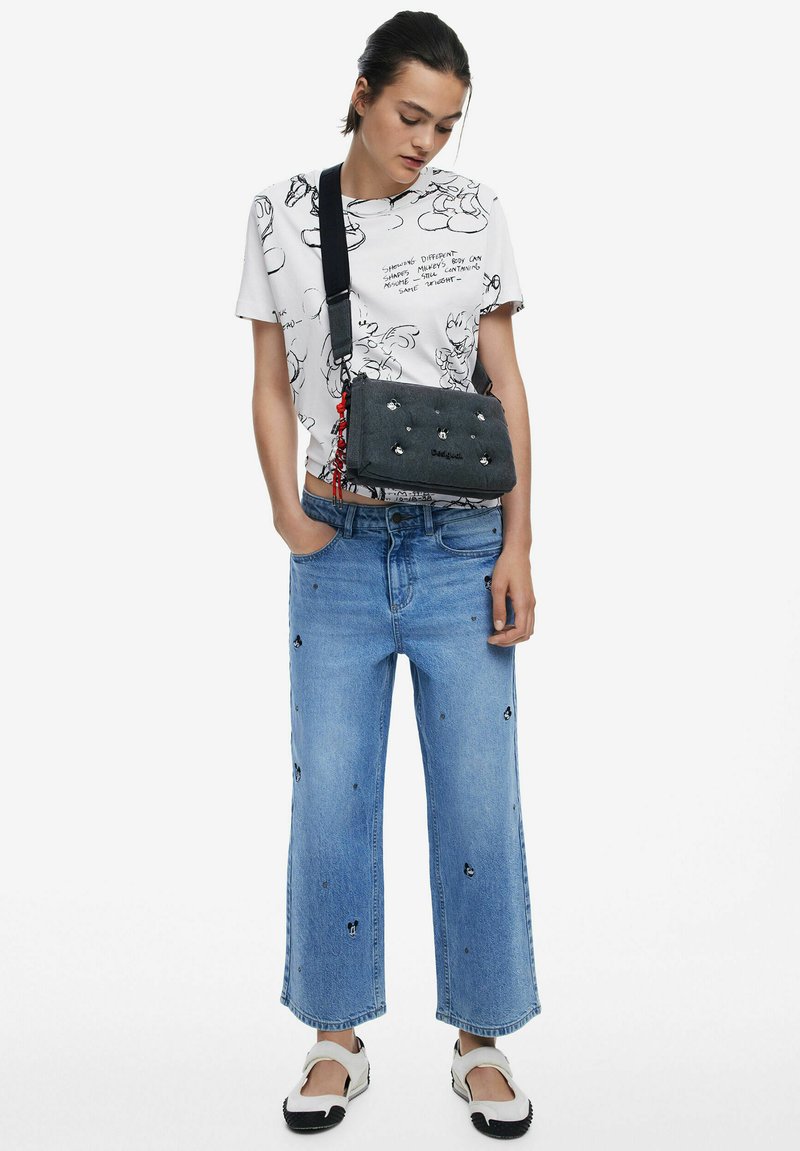 Graue Handtasche mit Verzierungen, weißes grafisches T-Shirt, hellblaue weit geschnittene Jeans mit Stickereien und schwarz-weiße Slip-On-Schuhe.