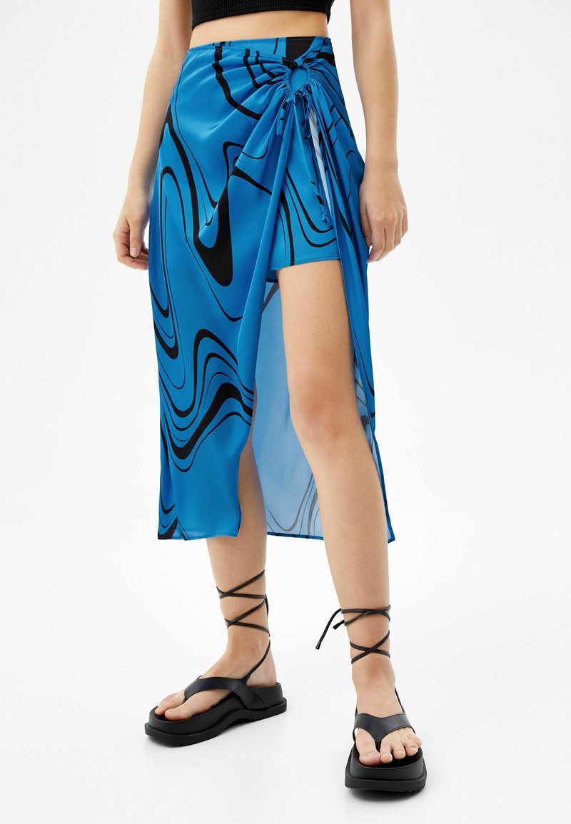 Sininen silkkiwrap-hame, jossa on abstrakti musta pyörteinen kuvio, korkea halkio ja solmittava yksityiskohta vyötäröllä, yhdistettynä mustiin litteisiin sandaaleihin.