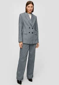 Grijs gestreepte, dubbelgeknoopte blazer met grote zwarte knopen, gedragen met bijpassende wijd-uitlopende broek. De stof heeft een gladde textuur.