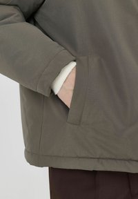 Veste vert olive foncé, avec une texture lisse, une poche latérale, un poignet en crème côtelé et un ourlet structuré.