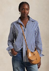 Vrouw die een blauw-wit verticaal gestreept overhemd draagt, blauwe spijkerbroek, hangende oorbellen en een beige leren crossbodytas, staat met één hand in de zak.