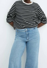 Personne portant une chemise à manches longues rayée noir et blanc et un jean large taille haute bleu clair, debout avec les mains dans le dos.