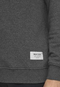 Grijze sweatshirt van gestructureerde stof, met een wit label met "INDICODE" en tekst. Ribgebreide boorden en zoom zichtbaar aan de onderkant.