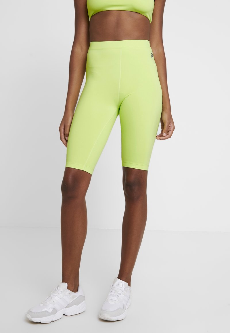 Cuissards de vélo vert néon à taille haute, texture lisse et coutures plates. Portés avec des chaussures de sport blanches, éléments de design complémentaires.