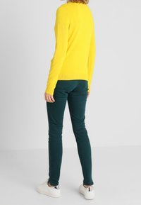 Žlutý svetr s dlouhým rukávem, který je spárován s tmavozelenými skinny džínami a bílými teniskami, přičemž boty mají stříbrné akcenty.