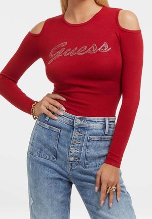 Femme portant un haut à manches longues rouge à épaules dénudées avec le texte "Guess" et un jean taille haute bleu avec des boutons à l'avant.