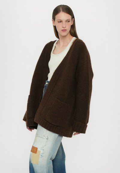 Brown oversized cardigan, textured fabric, open front, with two front pockets, worn over light-colored tank top and distressed jeans.