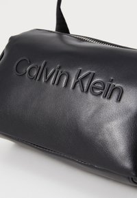 Črna usnjena torbica z vtisnjenim logotipom "Calvin Klein", ki ima teksturirano površino in kovinsko zadrgo za zapiranje. Kompaktna in pravokotna oblika.
