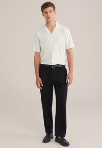 Camisa blanca de manga corta con cuello y cierre de botones, combinada con pantalones negros holgados y un cinturón negro trenzado. Zapatos negros sin cordones.