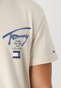 Beige bavlněné tričko s modrým logem obsahujícím názvy měst a malým červeným, bílým a modrým akcentem na rukávu.