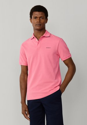 Man staat met hand in zak, gekleed in een roze Hackett polo en donkerblauwe korte broek tegen een effen grijze achtergrond.