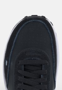 Černá teniska s kombinací texturovaných materiálů, zaoblenou špičkou, propracovaným vzorem tkaniček a bílou gumovou podrážkou s tmavými detaily.