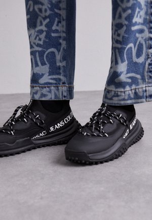 Schwarze Sneakers mit weißem "Jeans Couture" Text, getragen mit blauen Jeans mit Graffiti-Print auf grauem Fußboden-Hintergrund.