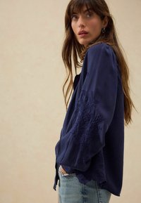 Blouse bleu marine avec des manches volumineuses ornées d'une riche broderie florale le long des poignets. Tissu doux, coupe décontractée, associée à un jean léger.