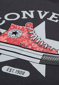 Raudonos Converse sportbačių grafika ant juodo fono, šalia baltos žvaigždės ir užrašo „CONVERSE EST. 1908.“ Sportbačiai turi raištelius ir išskirtinį raštą.