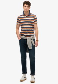 Polo w paski w granatowym, pomarańczowym i białym kolorze, zestawiony z ciemnymi dżinsami; bluza przewiązana w pasie, białe sneakersy z czarnymi akcentami.