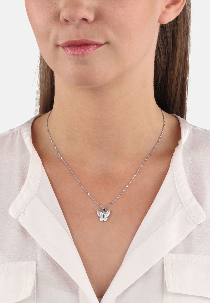 Donna che indossa una collana in argento con un piccolo pendente a forma di farfalla incastonato di pietre trasparenti, abbinata a una blusa bianca.