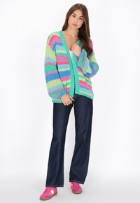 Mehrfarbiger Strickcardigan mit blauen, grünen, pinken und gelben Streifen; V-Ausschnitt, oversized Passform, kombiniert mit dunklen Jeans und pinken Turnschuhen.