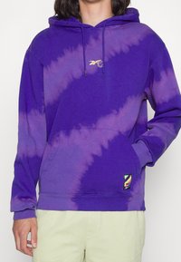 Violetti tie-dye huppari, jossa on etutasku, nyörihuppu ja pieni kultainen logo. Varustettu ribbaustuilla hihoilla ja helmalla, valmistettu pehmeästä kankaasta.