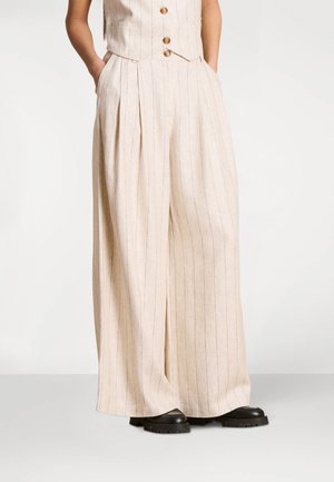 Femme portant un pantalon beige à jambes larges avec de fines rayures verticales et des chaussures noires épaisses, les mains dans les poches, debout sur un fond neutre.