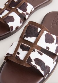 Braune Slide-Sandalen mit weißen und schwarzen Kuhfell-Riemen, die eine weiche, strukturierte Oberfläche und eine flache braune Sohle aufweisen.