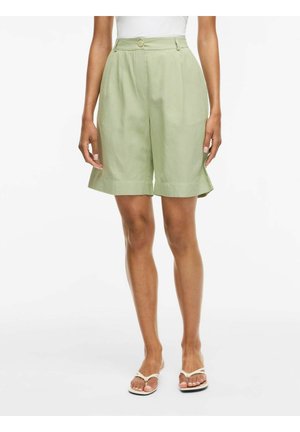 Vrouw draagt lichtgroene, hooggetailleerde knielange shorts met plooien, wit topje en beige teenslippers op een witte achtergrond.