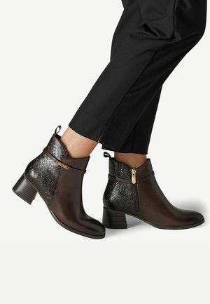 Bottes marron en cuir montantes à la cheville avec panneaux arrière texturés, fermeture éclair latérale, talons carrés, portées avec un pantalon noir raccourci.
