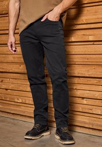 Czarne, slim-fit spodnie wykonane z bawełny o gładkiej strukturze. Z przodu kieszenie i swobodny krój. Połączone z ciemnymi butami na tle drewna.