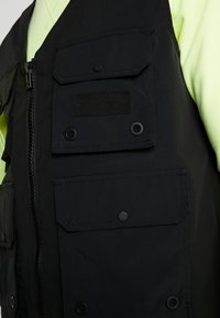 Svart väst i ett slätt tyg med flera framfickor, tryckknappsstängning och en central dragkedja. Bärs över en neongrön topp.