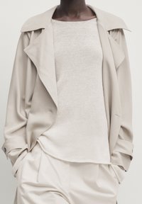 Trench-coat beige avec col large, tissu léger et coupe décontractée portée sur un haut en maille légère. Associé à un pantalon crème avec une texture subtile.