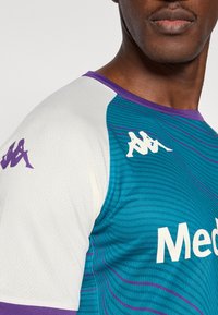 Športna majica s kratkimi rokavi v barvi zeleno-modre z vijoličnimi poudarki, tkanina z drobnim mrežastim vzorcem, na sprednji strani bel logotip »Med« in logotip na rokavih.