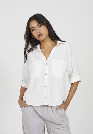 Giovane donna con lunghi capelli scuri che indossa una camicia bianca con maniche arrotolate e pantaloni grigio chiaro, mani nelle tasche, sfondo neutro.