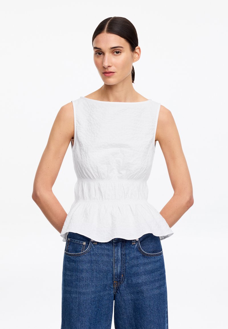 Femme aux cheveux foncés attachés porte un top blanc sans manches à basque et un jean bleu taille haute, debout devant un fond blanc uni.