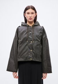 Õunaroheline lühike jope kapuutsiga, millel on üle suurendatud varrukad, trukk-nupud ja pruunid nöörid reguleerimiseks. Sile, mati tekstuuriga.