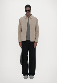 Beige jacket with two chest pockets, gray shirt underneath, black wide-leg pants, gray sneakers, and black bag, worn by a model with sunglasses.