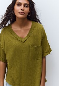 Olivengroene V-hals T-shirt gemaakt van een zachte, gestructureerde stof, met opgerolde korte mouwen en een zak aan de linkerkant.