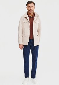Jasno beżowa kurtka z kapturem, z dwiema przednimi kieszeniami, noszona na ciemnobrązowej koszulce polo, w połączeniu z ciemnoniebieskimi dżinsami i białymi trampkami.