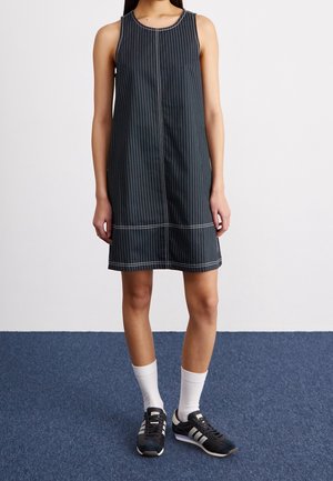 Nainen pukeutuneena mustaan, hihattomaan puvuntakkiin, valkoisiin nilkkasukkiin ja mustiin Adidas-tossuihin seisomassa sinisellä matolla.