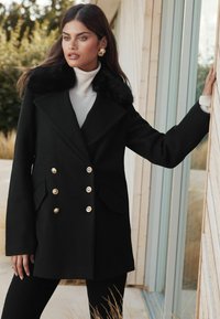 Čierny dvojradový kabát so zlatými gombíkmi a golierom z umelej kožušiny, v kombinácii s bielym rolákom a čiernymi nohavicami. Hladká textúra.