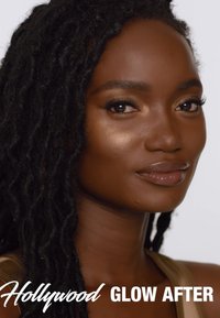 Une mannequin avec de longs cheveux ondulés et noirs et une peau lisse pose dans un haut beige. Un maquillage naturel met en valeur ses traits sur un fond blanc.