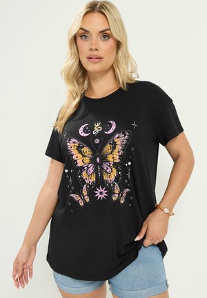 Kvinde med blond hår har en sort T-shirt med et stort pink og gult sommerfugle- og halvmåne-design, kombineret med lyseblå shorts.