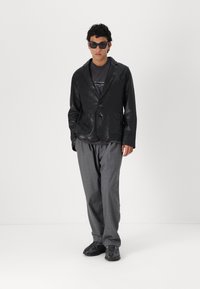 Schwarze Lederblazer mit aufgesetzten Taschen, getragen über einem grauen T-Shirt, kombiniert mit grauen Hosen und schwarzen Sneakers. Das Model trägt dunkle Sonnenbrillen.