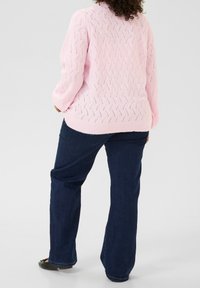 Lyserød strikket trøje med et struktureret mønster, parret med mørkeblå lige jeans og sorte flade sko. Fokuseret på bagvæg.
