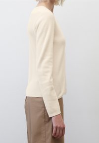 Pull texturé couleur crème à manches longues et col rond, associé à un pantalon beige, vu de profil sur un fond uni.