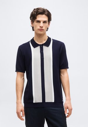 Νέος άνδρας με κυματιστά μαλλιά, φορώντας μπλε πόλο με κάθετες γκρι και λευκές ρίγες, στέκεται μπροστά σε απλό φωτεινό φόντο.