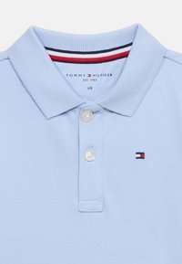 Ανοιχτό μπλε πόλο πουκάμισο από υφασμάτινη υφή, με μυτερό γιακά, τρία κουμπιά και μικρό κεντημένο λογότυπο σε κόκκινο, άσπρο και ναυτικό μπλε.