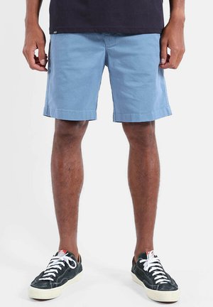Homme portant un short bleu clair, des baskets noires avec des lacets blancs, et une chemise noire, debout les mains près des poches du short.