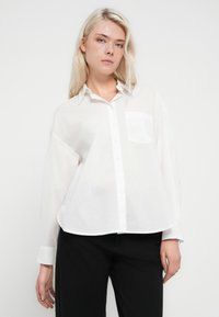 Bela srajca z gumbi in žepom, izdelana iz lahkega blaga, s klasičnim ovratnikom in sproščenim krojem. Združena s črnimi hlačami.