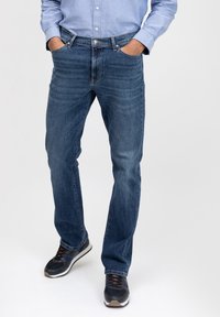 Vaqueros de mezclilla azul con un diseño de pierna recta, con un ligero desgastado, cinco bolsillos y costuras en contraste. Combinados con zapatillas oscuras.