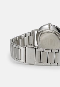 Reloj de acero inoxidable con acabado pulido, que cuenta con una pulsera de eslabones y un logo grabado en el cierre. Caja redonda con un fondo liso.